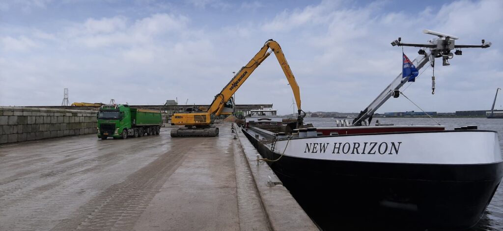 Port scene with a green dump truck and a yellow excavator on a wet dock beside a boat named New Horizon being loaded by a crane.